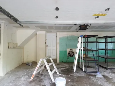A garage interior showing a painting project in progress. The ceiling is painted white, while walls are a mix of light beige, unpainted drywall with mudding, and an old peeling green wall. Two ladders, a white paint bucket, and metal shelving are visible on the concrete floor.