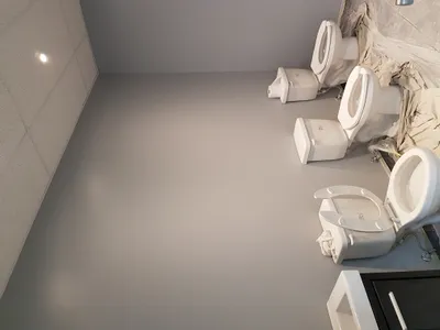 View of a commercial restroom with multiple white toilets protected by plastic and drop cloths. The walls are painted a smooth, light gray, contrasting with the white drop ceiling on the left. The image showcases the completed wall painting work.