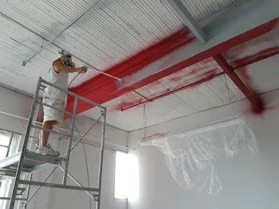 A painter on scaffolding spray painting a large commercial ceiling. Red paint is being applied to exposed metal beams and parts of the white plank ceiling. The painter wears a respirator and white work clothes. The walls are white brick, and a light fixture is covered in plastic.