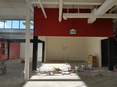 Photo taken during the painting of a commercial interior, showing a large open space with a newly painted bold red accent wall and light beige walls. The exposed ceiling, ductwork, and structural columns are painted white and black respectively. Paint cans, drop cloths, and construction equipment are visible on the concrete floor, indicating work in progress.