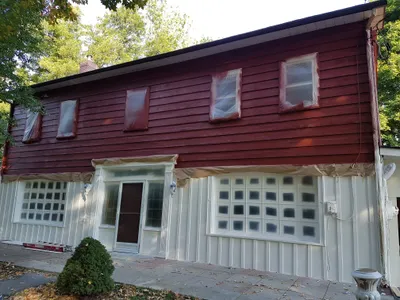 Exterior view of a residential house undergoing painting. The upper siding is a reddish-brown, while the lower siding and trim are freshly painted white. Windows are masked with plastic and paper for protection, indicating work in progress on the house facade.