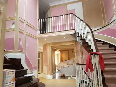 A large residential foyer and grand staircase during a painting project. Walls with wainscoting are visible, showing patches of white primer over a pink base coat, indicating drywall repair and prep work. The dark wood staircase with white balusters and a light pink carpet runner is prominent. Boxes of drywall compound are on the stairs, and floors are covered with protective paper, signifying an active painting job.