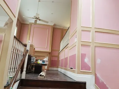 Interior view of a residential foyer and staircase during a painting project. Walls feature decorative paneling, with pink sections and beige trim showing primer and spackle patches. White balusters and baseboards are visible, along with a dark wood staircase and a white ceiling with a fan.