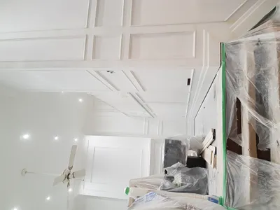 A residential interior painting project in progress, featuring high white walls with intricate decorative paneling and trim. The white ceiling with recessed lighting and a ceiling fan is also visible. Furniture and flooring are covered with plastic sheeting and green painter's tape.