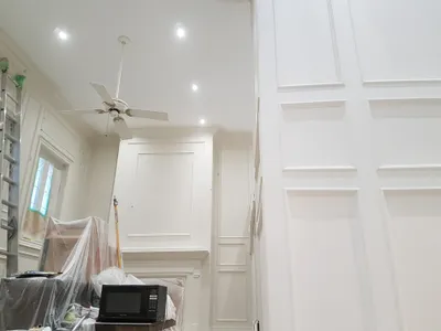 Interior view of a residential living room or foyer during a painting project, featuring freshly painted white walls with decorative paneling, white ceiling, and white trim around a window and fireplace. A ladder and covered furniture indicate work in progress.