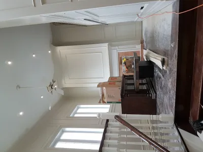 Interior view of a residential living room or foyer during a painting project, showcasing freshly painted white wall panels, crown molding, and ceiling. A staircase with white balusters and a dark wood handrail is visible. The floor is dusty, and moving boxes are present, indicating the 'during' phase of the project.