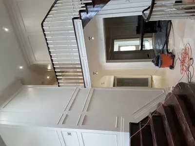 A residential foyer and staircase with newly painted white wainscoting, trim, and balusters. The walls are a light neutral color, and the stair treads and handrail are dark wood. A vacuum cleaner and extension cord are visible on the floor, indicating ongoing work.