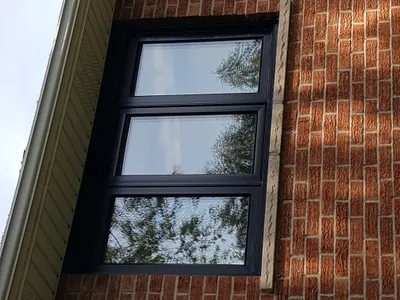 Close-up of a building exterior showcasing newly painted black window frames. The windows are set into a textured red brick wall, with reflections of trees and a bright sky. A light-colored gutter and soffit are visible on the left side of the image.