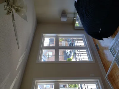 A bedroom with light tan painted walls and crisp white window trim. A large window looks out onto a residential street. A white popcorn ceiling and a ceiling fan are visible. The room features a bed with dark bedding and a modern lamp.
