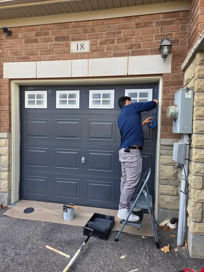 A painter on a step ladder applies blue painter's tape around the trim of a garage door. The garage door is being painted a dark gray color, with paint rollers and trays visible on the driveway. The house exterior features brick and stone accents.