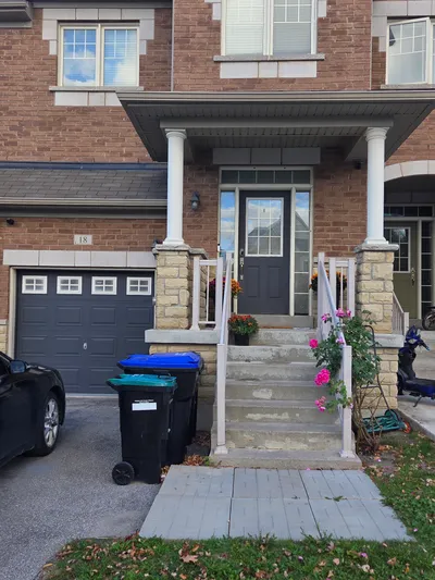 Front exterior of a brick townhouse featuring a dark gray painted front door and matching garage door. White painted porch columns and railings frame the entrance. The house has light-colored window trim and a paved driveway with a black car and garbage bins.