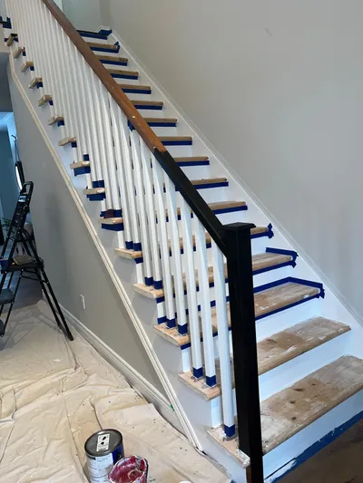 A residential staircase during a painting project. White balusters and risers are visible, with blue painter's tape protecting the wooden treads and parts of the handrail. The newel post and a section of the handrail are painted black. Drop cloths cover the floor, and paint cans are visible, indicating active work.
