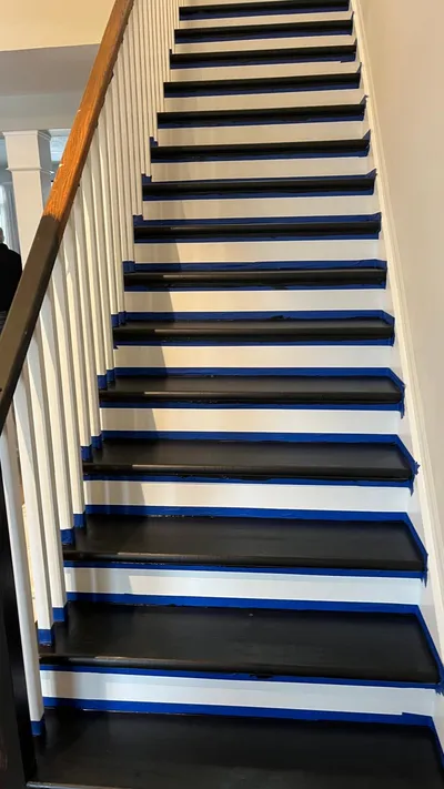 View of a residential staircase during a painting project. The stair treads are freshly painted black, while the risers are white. Blue painter's tape is visible along the edges of the treads and risers, indicating work in progress. A wooden handrail and white balusters are also visible on the left.