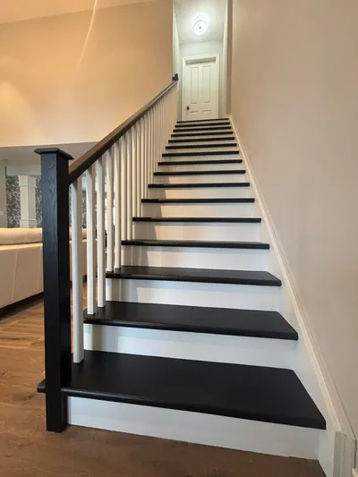 A newly painted modern staircase featuring dark black treads, crisp white risers, white balusters, and a dark newel post and handrail. The surrounding walls are painted in a light, neutral color, showcasing a clean and updated look.