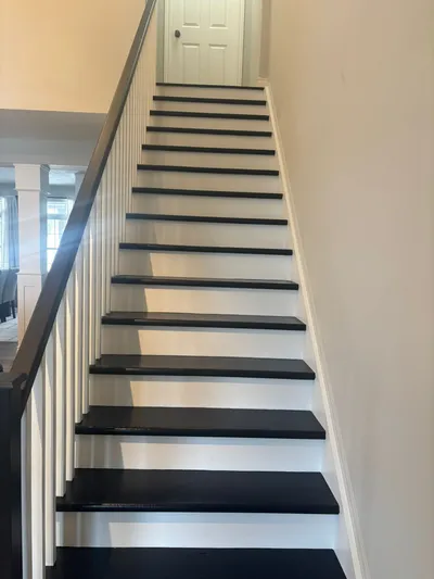 A newly painted staircase featuring dark black treads and crisp white risers, balusters, and trim. The handrail is painted a dark brown or black, complementing the treads. The surrounding walls are painted a light, neutral beige or off-white color. The photo showcases a clean, finished look for residential interior painting.