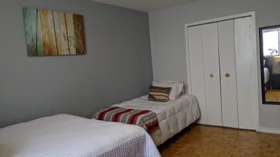 A residential bedroom featuring walls painted in a contemporary gray, complemented by crisp white trim and closet doors. The room contains two beds with white bedding, a patterned throw, and a parquet wood floor. A piece of abstract art hangs on the gray wall.
