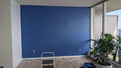 A room featuring a newly painted bold blue accent wall, with white walls and ceiling. Drop cloths cover the floor, and a step ladder, paint tray, and roller are visible, indicating the painting project is in progress. Large windows with a view are on the right.