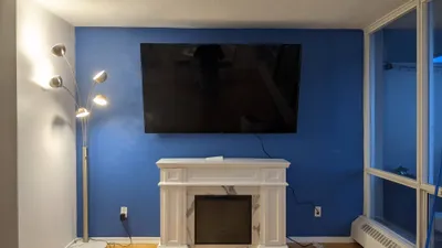 A modern living room featuring a vibrant blue accent wall with a mounted television above a white decorative fireplace. The adjacent wall is painted a light gray, and a multi-head floor lamp stands in the corner. A large window is visible on the right, showing a glimpse of the exterior.