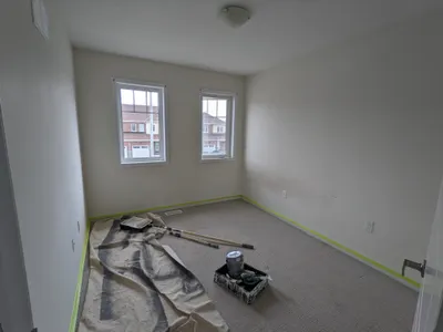 Interior view of a residential bedroom during a painting project. Walls are light beige, with some areas appearing freshly painted white. Painter's tape lines the baseboards, and a drop cloth covers part of the carpeted floor. Paint cans, rollers, and brushes are visible on the floor.
