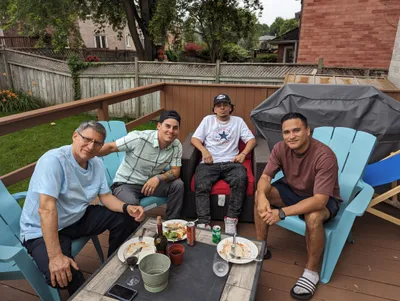 Four men sitting around a table on a brown stained wooden deck with matching railings, enjoying food and drinks outdoors. The deck appears to be recently stained.