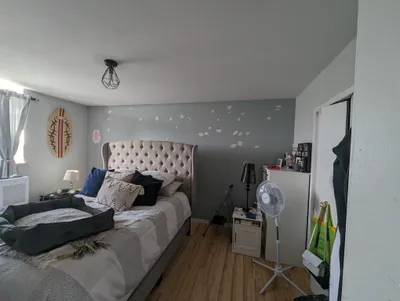 A bedroom showing a gray accent wall behind a bed with visible white drywall patches, indicating preparation for a new paint job. The ceiling is white, and the room contains a bed with a tufted headboard, nightstands, and a fan.