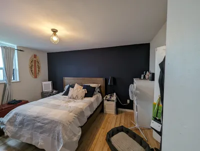 A finished bedroom featuring a dark navy or charcoal accent wall behind a wooden bed frame, contrasted with light off-white painted walls and a white ceiling. The room is furnished with a bed, nightstands, and a window with curtains.