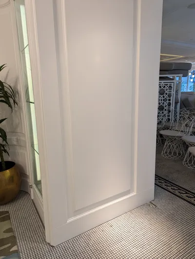 A close-up view of freshly painted white wall paneling and trim in a commercial dining or lounge area, showcasing clean lines and a bright finish. A glass display case and a plant in a gold pot are visible on the left, with modern white chairs and patterned floor tiles in the background.