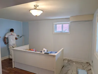 A painter wearing a Benjamin Moore shirt uses a roller to apply light blue paint to a bedroom wall. The room features a white crib, hardwood floors, and a window, with drop cloths protecting the floor during the painting process.
