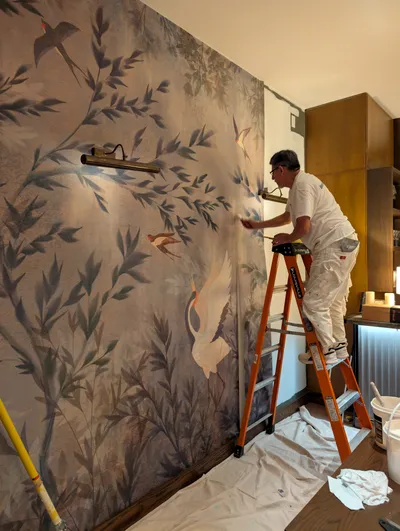 A painter in white overalls stands on an orange ladder, applying a finish with a brush to a large accent wall. The wall features a hand-painted mural with birds and botanical elements in muted greens, browns, and creams. Drop cloths are on the floor, and wooden cabinets are visible in the background, indicating an interior residential painting project.