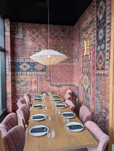 A commercial dining room featuring a long wooden table set with white plates and black napkins, surrounded by plush pink chairs. The walls are adorned with a rich, multi-patterned wallpaper resembling antique rugs in shades of red, blue, orange, and beige. The ceiling is painted a sleek matte black, contrasting with the vibrant walls. A large, white, pleated pendant light hangs above the table, and a gold wall sconce is visible on the right wall.