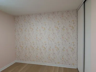A finished bedroom featuring a light pink painted wall, a contrasting accent wall with delicate floral wallpaper in pastel shades, and crisp white painted baseboards. The ceiling is white with a popcorn texture, and there are white sliding closet doors on the right. The floor is light wood laminate.