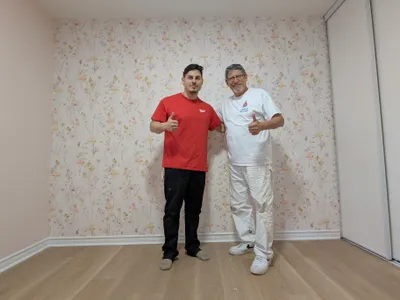 Two painters giving thumbs up in a newly finished residential room, featuring a light pink painted wall, crisp white baseboards, and a beautiful floral wallpaper accent wall with pastel flowers and butterflies. The room has light wood flooring and white sliding closet doors.