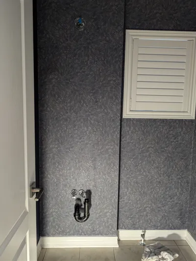 A bathroom wall featuring dark gray patterned wallpaper, with white painted window trim, white baseboards, and a white painted door frame. Exposed plumbing for a sink is visible on the wall.