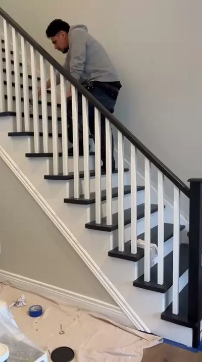A painter in a gray hoodie and jeans meticulously paints white balusters on a staircase. The staircase features contrasting black treads and handrail, white risers, and light gray walls. Drop cloths and painting supplies are visible on the floor, indicating an active painting project.