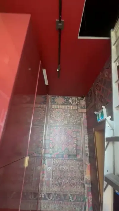 A rotated image of a commercial hallway featuring a vibrant red painted ceiling. The walls are adorned with intricate, colorful patterned wallpaper. A black track lighting system is installed on the ceiling, and a green exit sign is visible on the right wall.